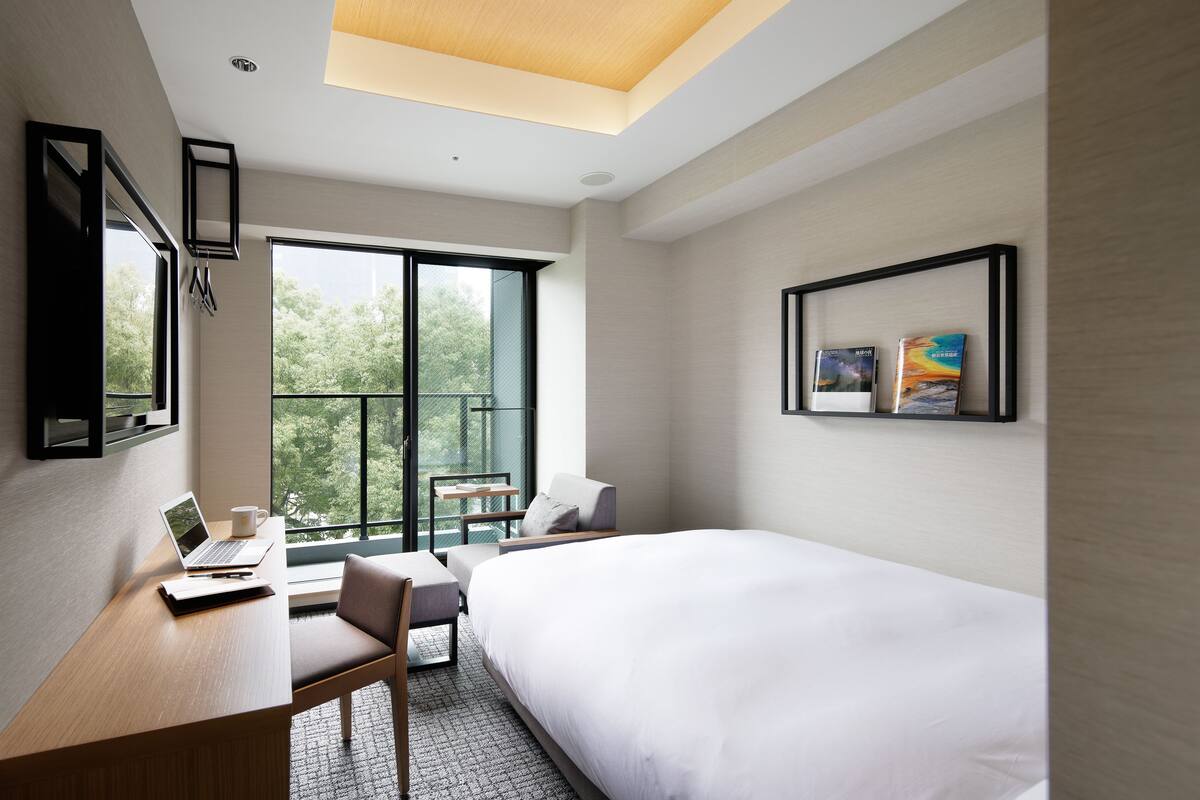 Una habitación de hotel moderna con una cama grande, un escritorio con una computadora portátil, una silla y un balcón con vista a árboles.