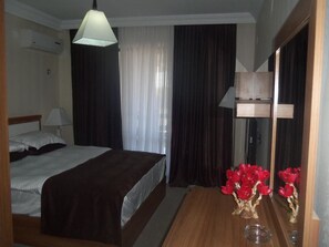 A hotel room with a bed, a wooden dresser, a mirror, and a vase with red flowers.