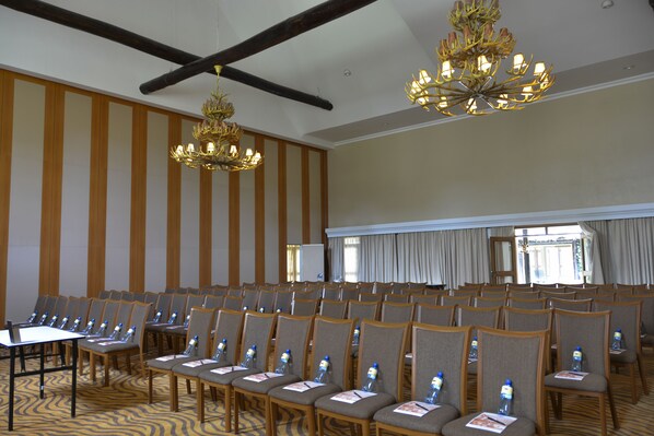 Uma sala de conferências com fileiras de cadeiras, uma mesa com um laptop e duas luminárias de cristal.