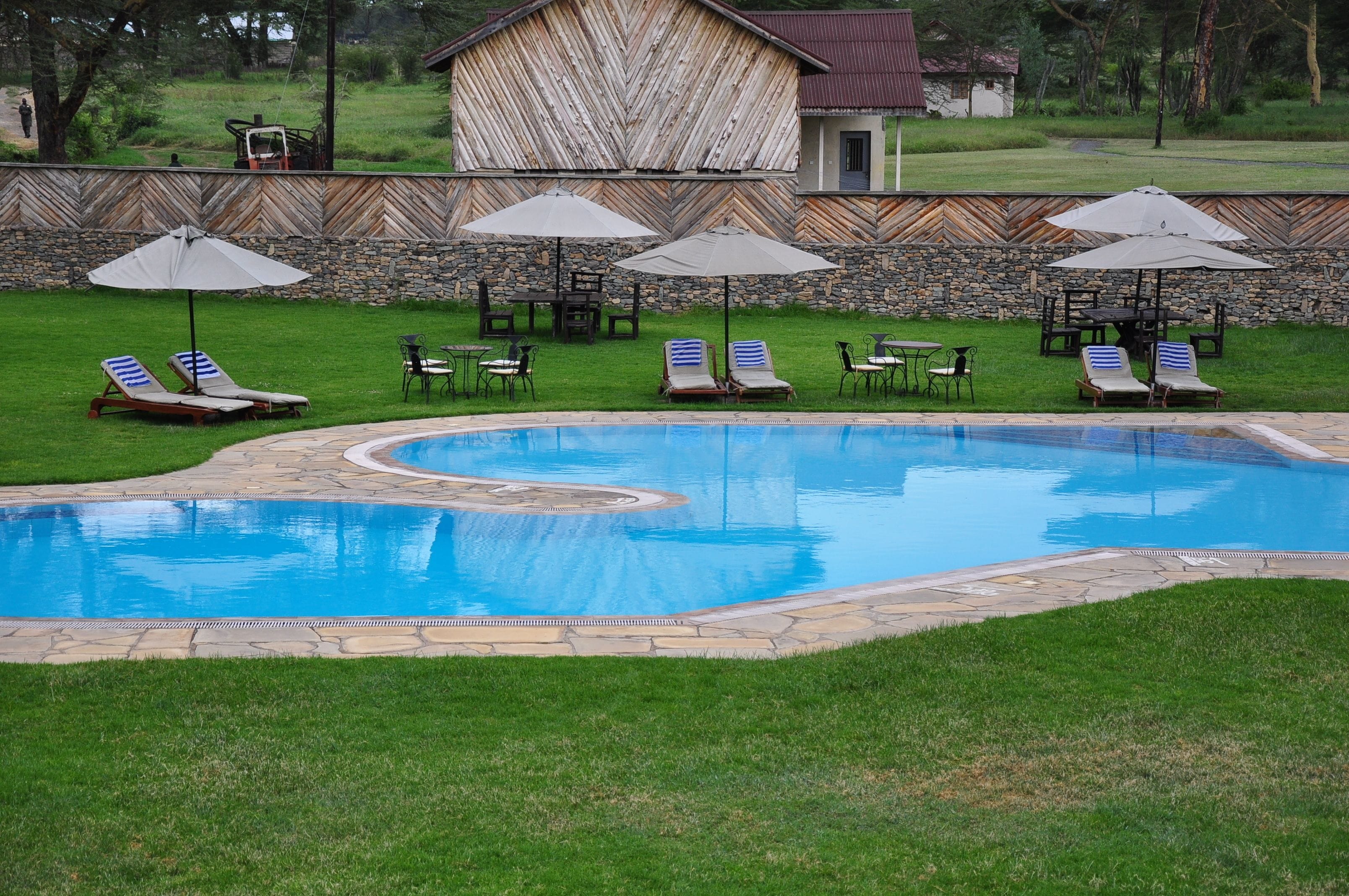Piscina com espreguiçadeiras e guarda-sóis, cercada por um muro de pedra e um edifício de madeira.