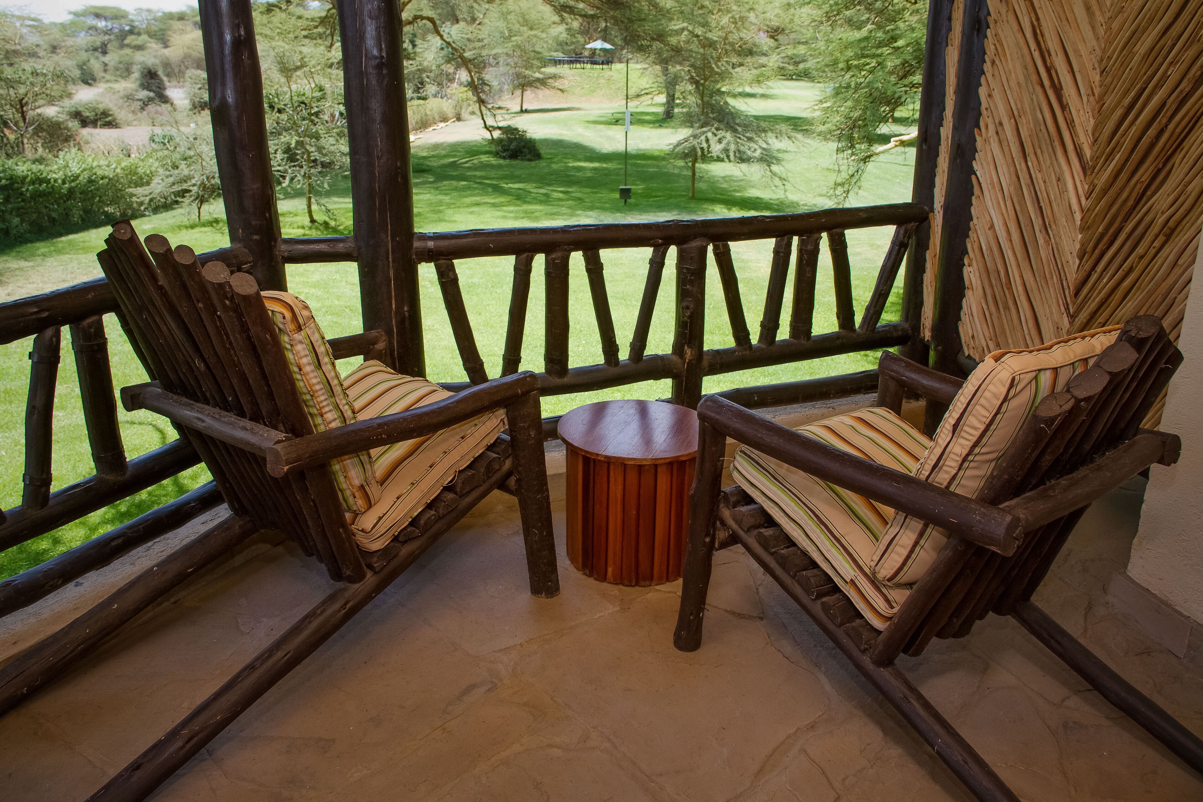 Une terrasse en bois avec deux chaises et une petite table, donnant sur une zone herbeuse bordée d’arbres et sur un bâtiment situé au loin.