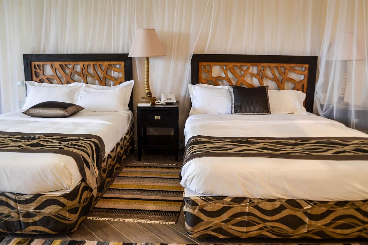 Two double beds with patterned bedspreads, a nightstand with a lamp, and a woven rug.