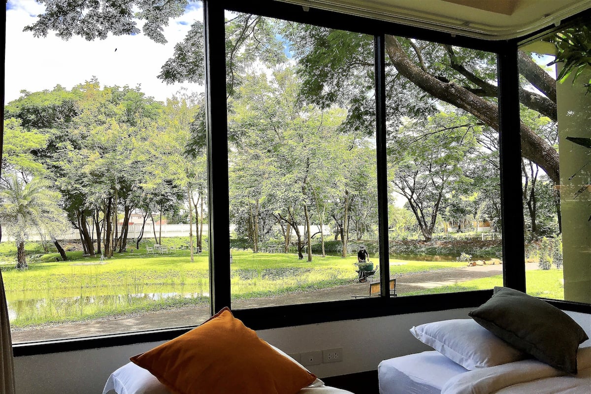 Quarto com uma janela grande que oferece vista para um parque com árvores e lago. Há duas camas com almofadas.
