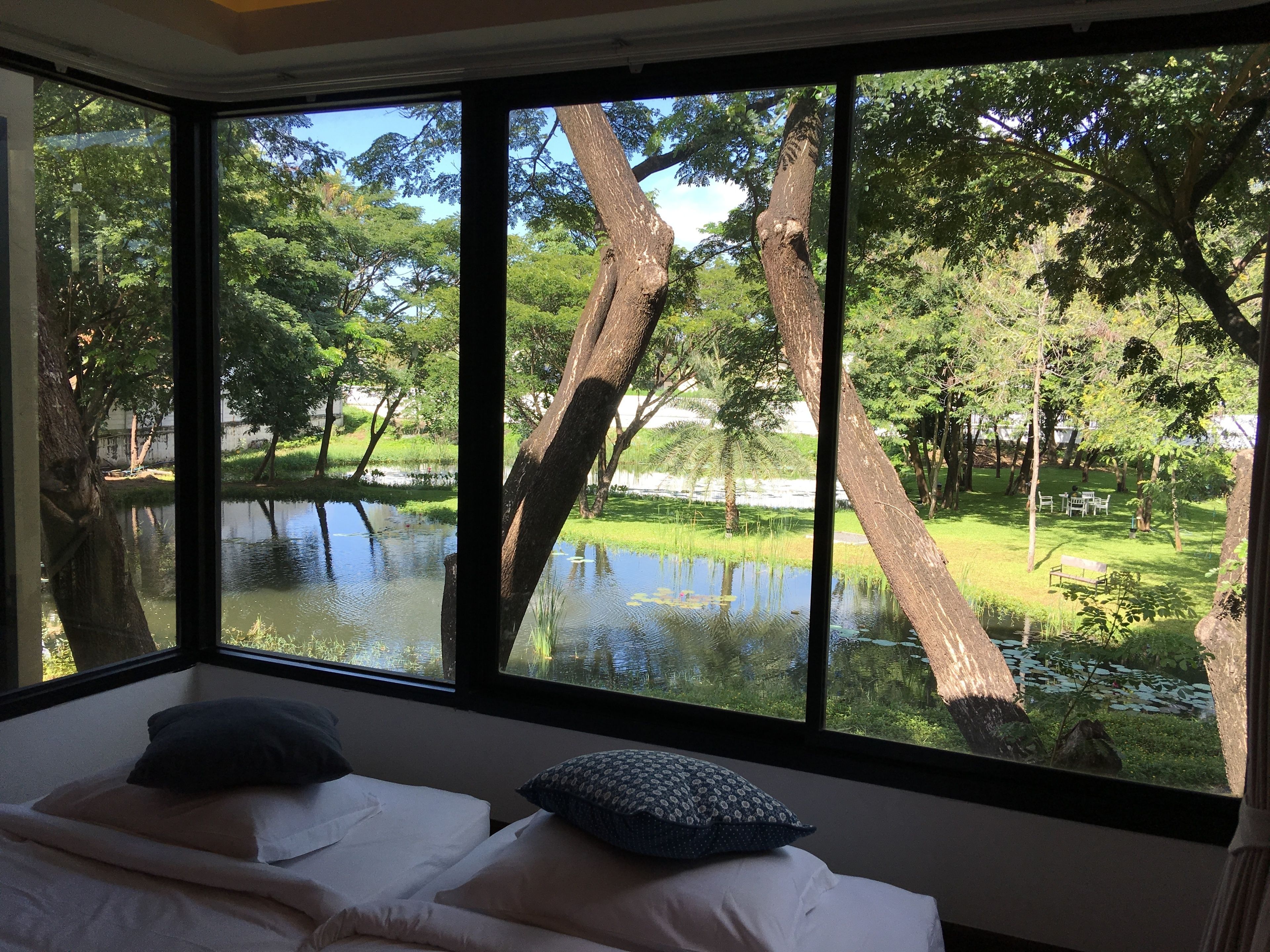 A room with a large window overlooking a pond and trees. There are two beds with pillows.