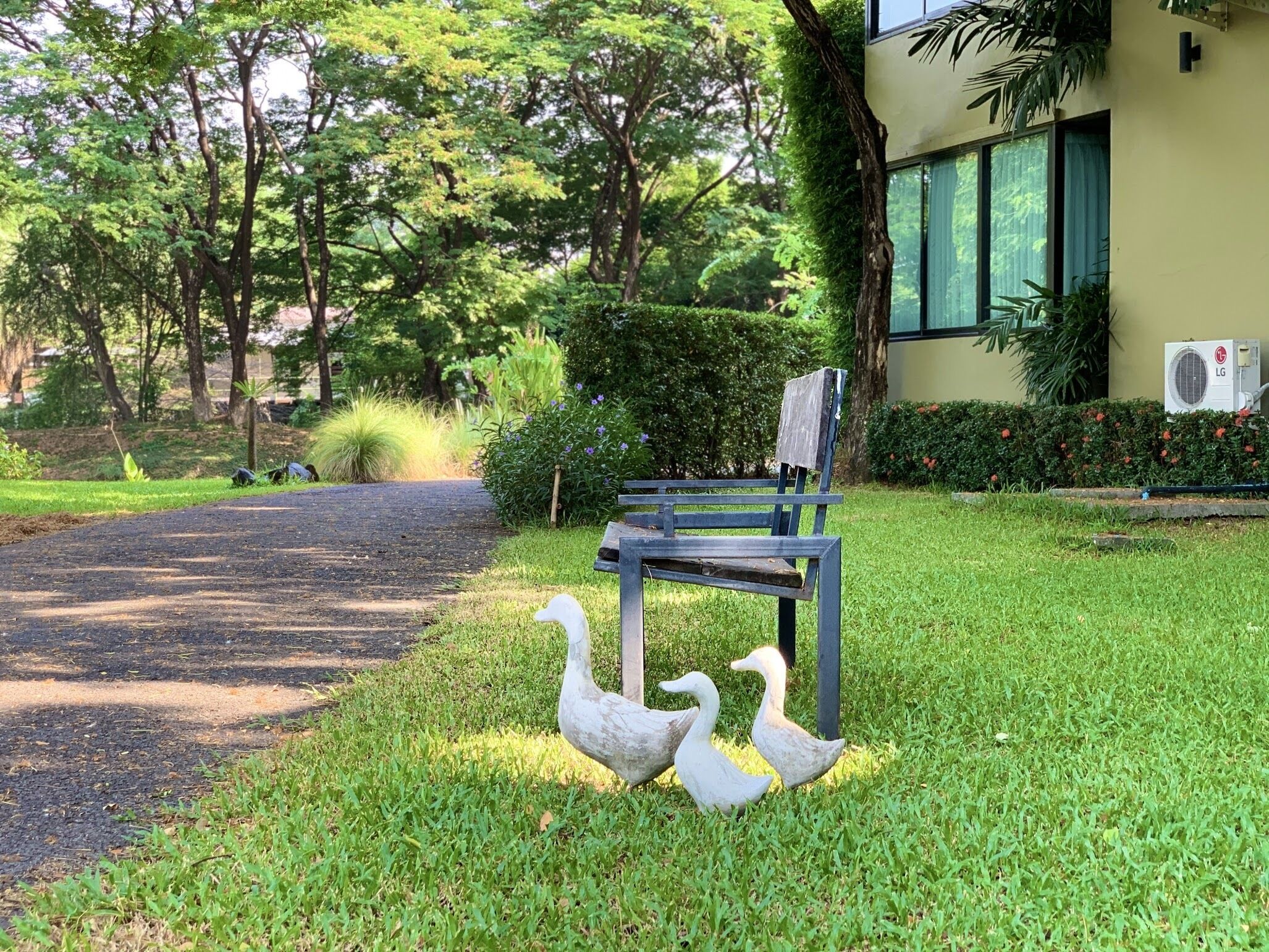 Un banco junto a una silla, dos esculturas de cisnes y un sendero que conduce a un edificio con puerta verde y un equipo de aire acondicionado.