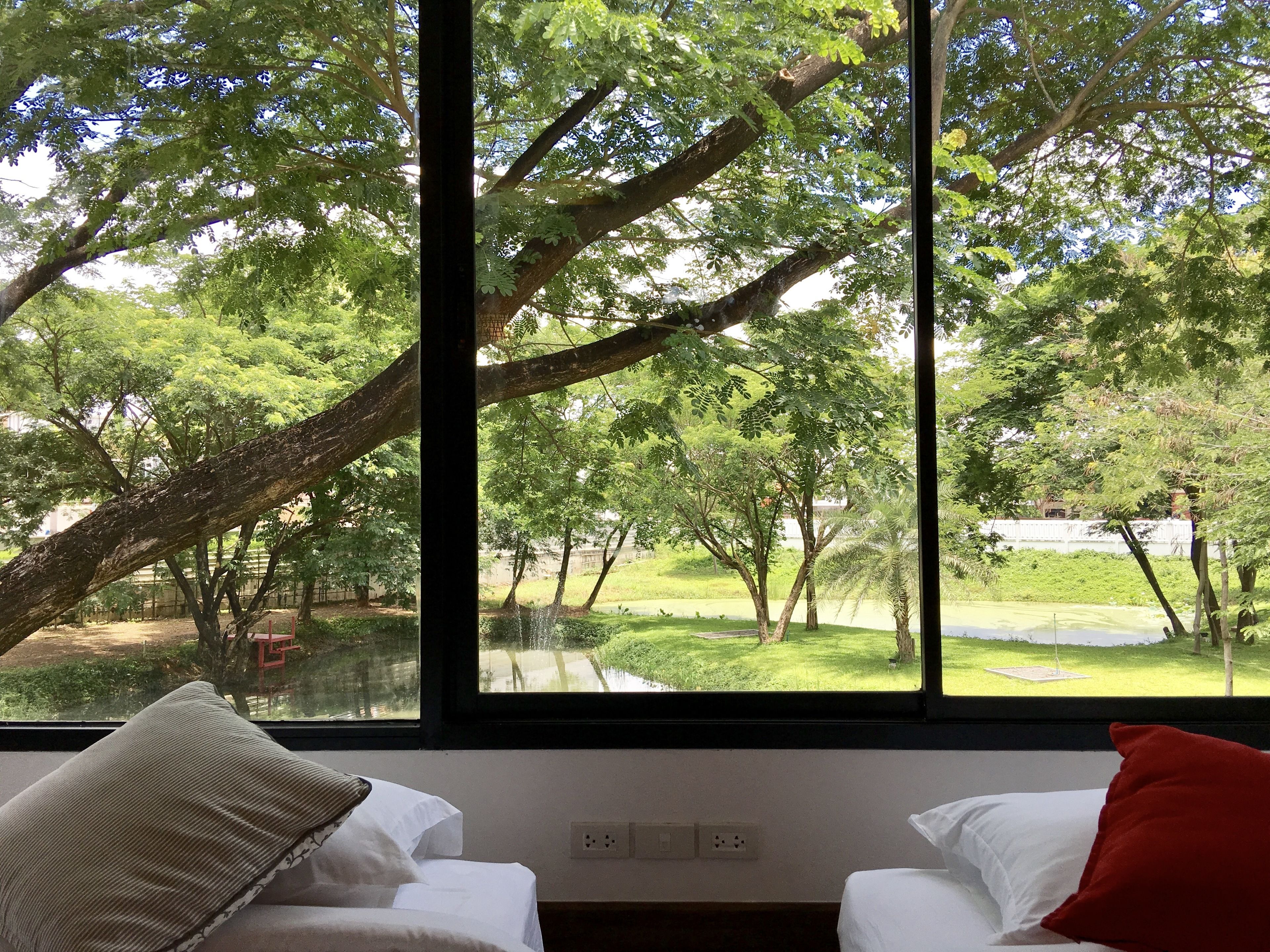 A room with a large window overlooking a lush green landscape with trees and a pathway. There are two beds with white bedding and a red pillow on one bed.