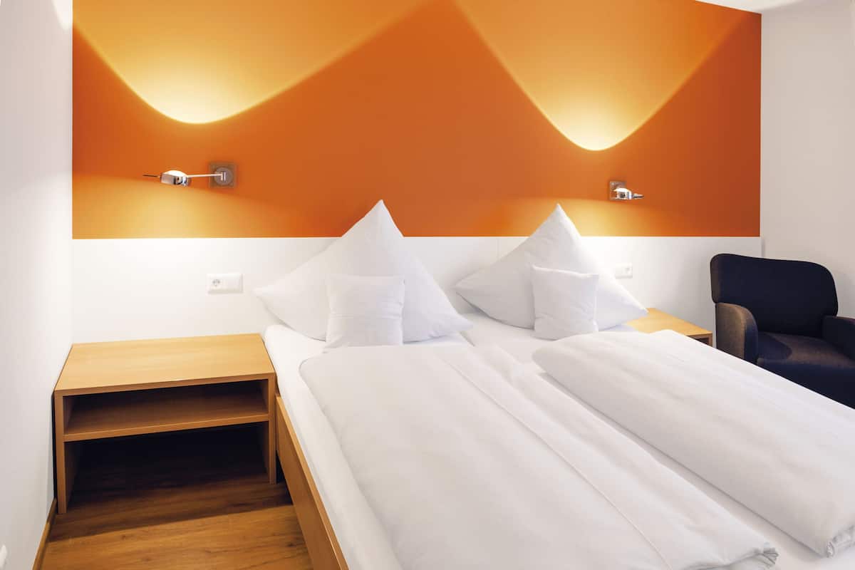 Una habitación de hotel moderna con una cama, almohadas, una mesita de noche, una silla y una lámpara fijada a la pared.