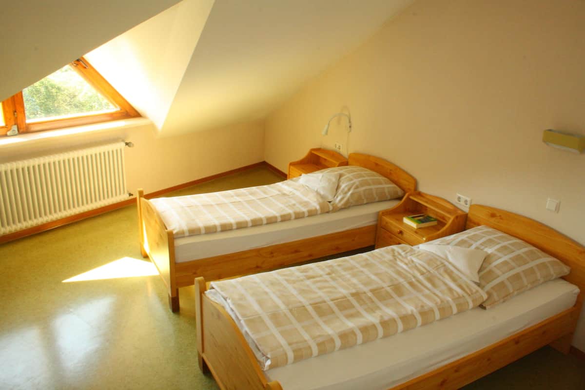 Una habitación pequeña y bien iluminada con dos camas de madera, una ventana que deja entrar luz natural y un radiador en la pared.
