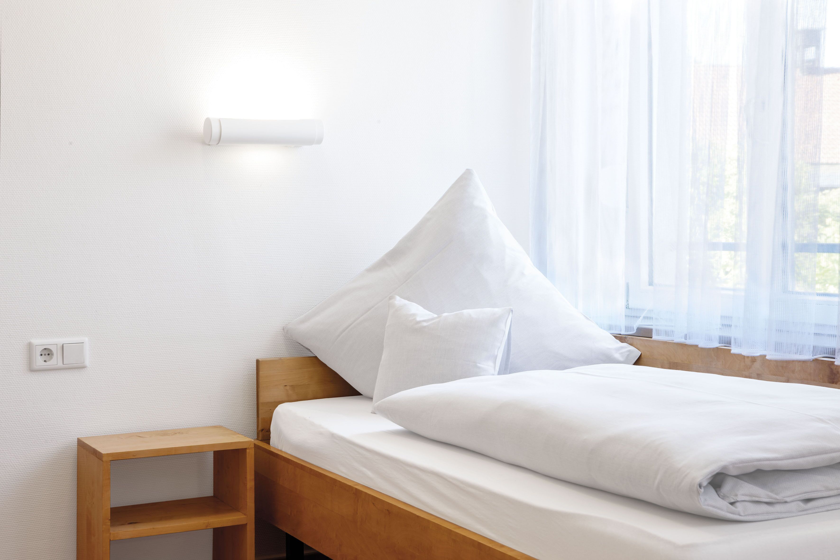 Un lit bien fait, avec du linge de lit blanc et des oreillers, une table de chevet en bois et une prise électrique au mur.
