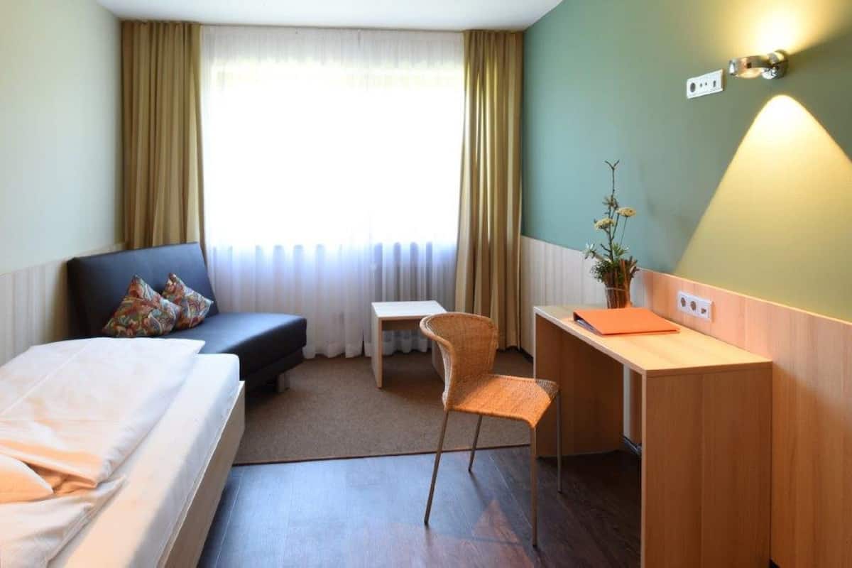 Una habitación de hotel con cama, escritorio, silla, mesita y ventana con cortinas.