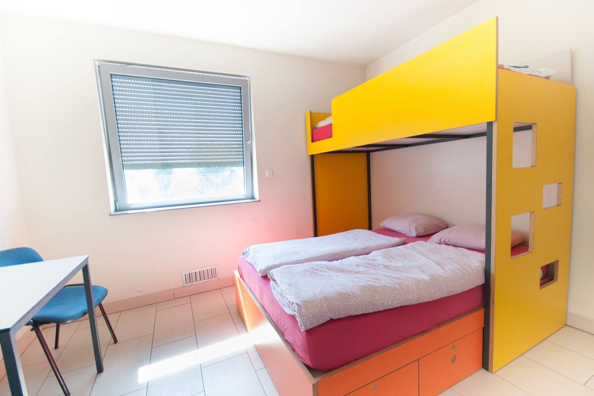 Uma cama de beliche com beliche superior amarelo e beliche inferior rosa. Há uma janela com persianas, uma secretária com cadeira e um radiador no chão.