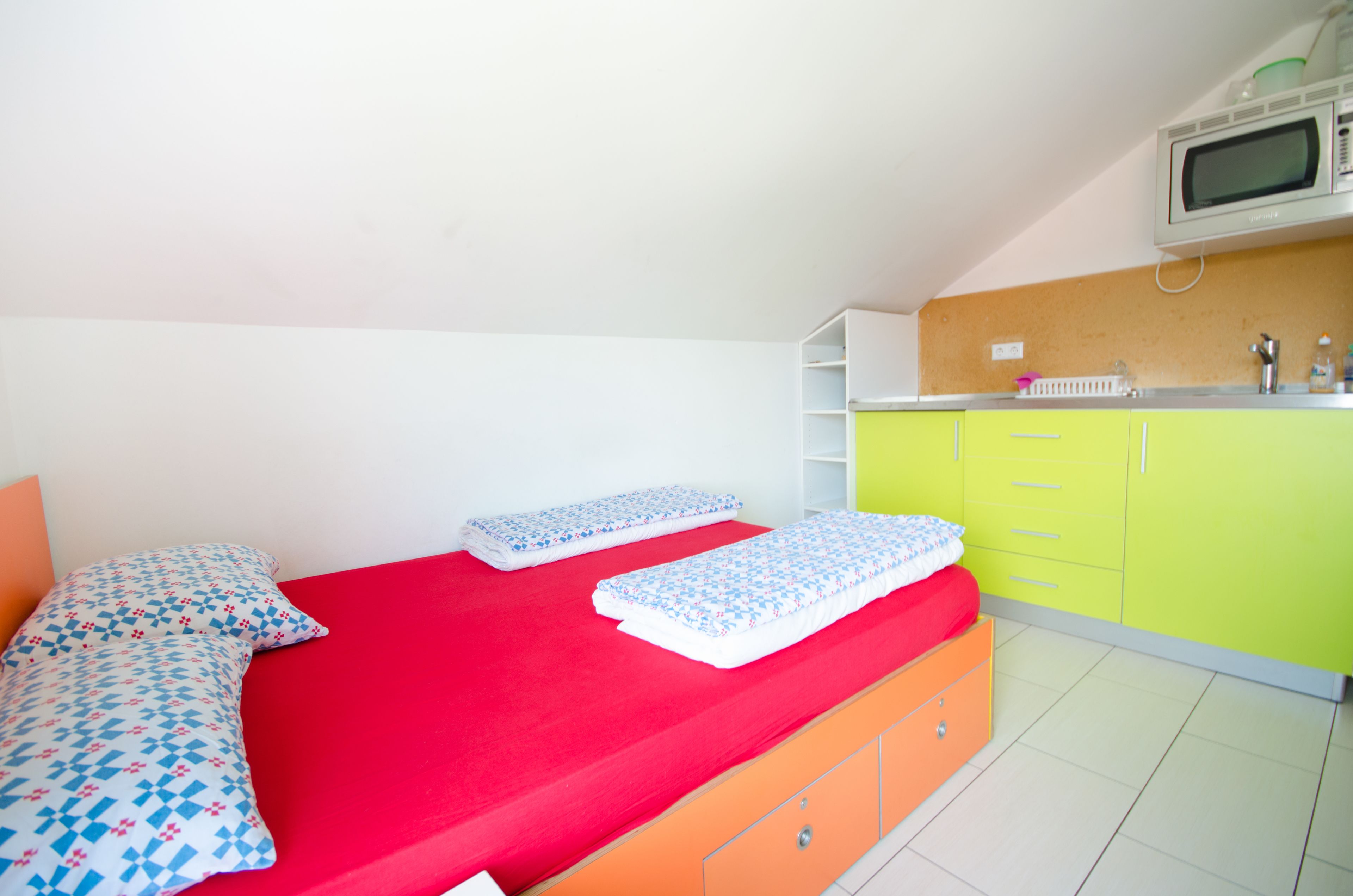 A small, bright room with a red bed, orange drawers, and a green cabinet.