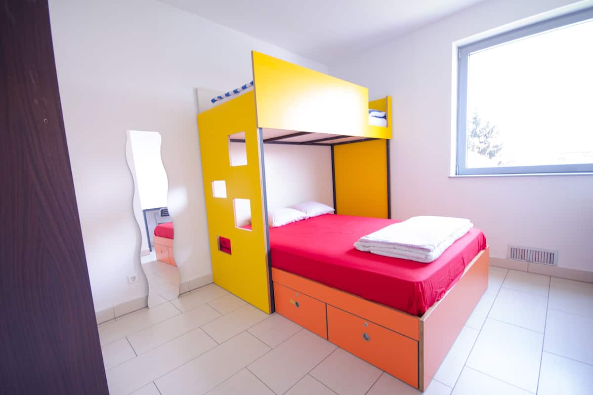 Uma cama beliche com colchão vermelho e cabeceira amarela, um radiador branco, e uma janela com vista para árvores.