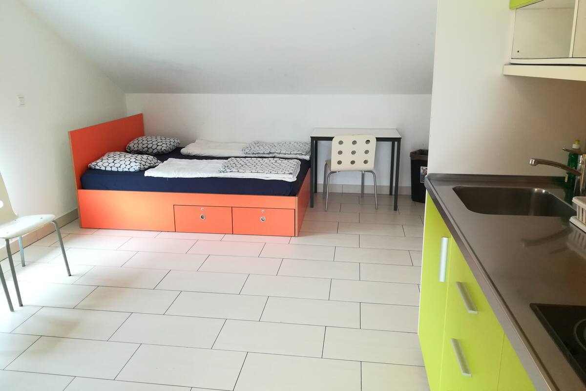 Quarto compacto com cama, uma mesinha, uma cadeira e um espaço com pia.