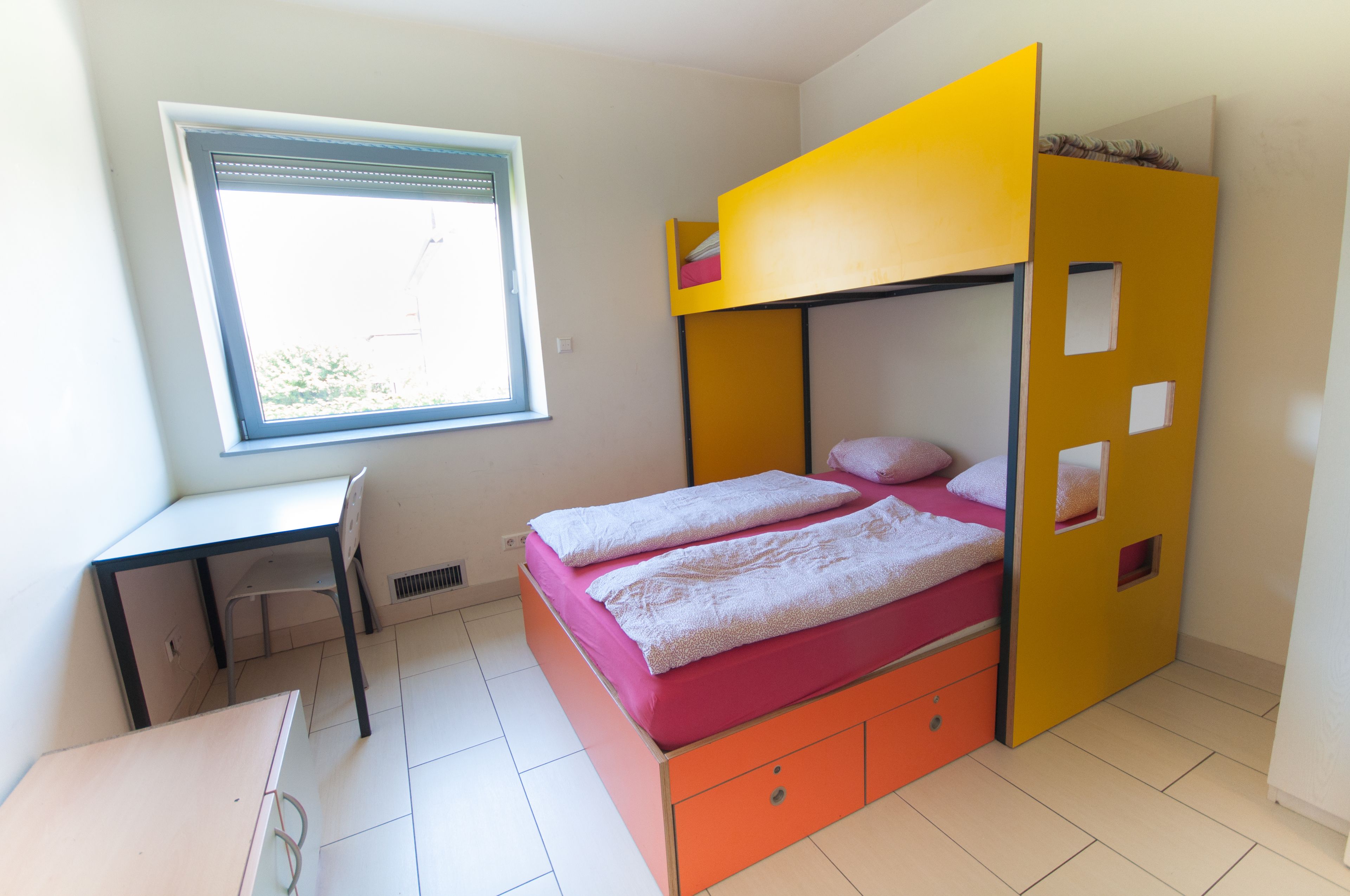 A bunk bed with a desk and chair, a window, and a tiled floor.