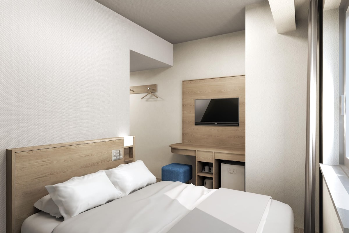 A modern hotel room with a wooden headboard, a flat-screen TV mounted on the wall, a desk with a chair, and a window providing a view.