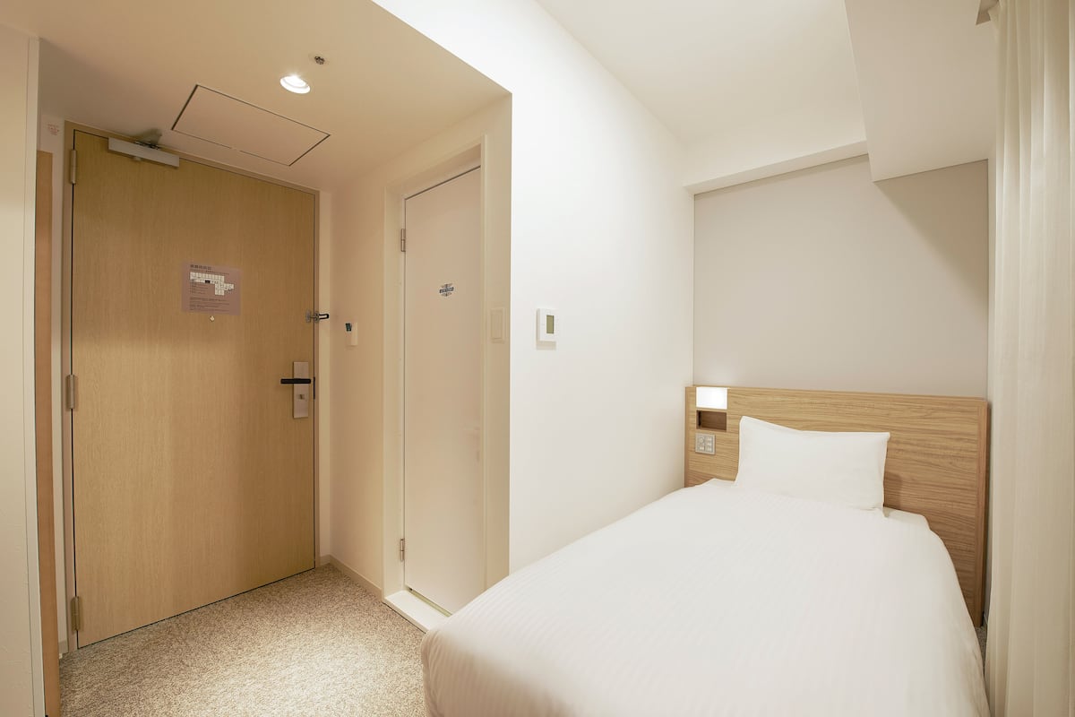 A hotel room with a wooden door, a bed with a wooden headboard, and a nightstand with a lamp.
