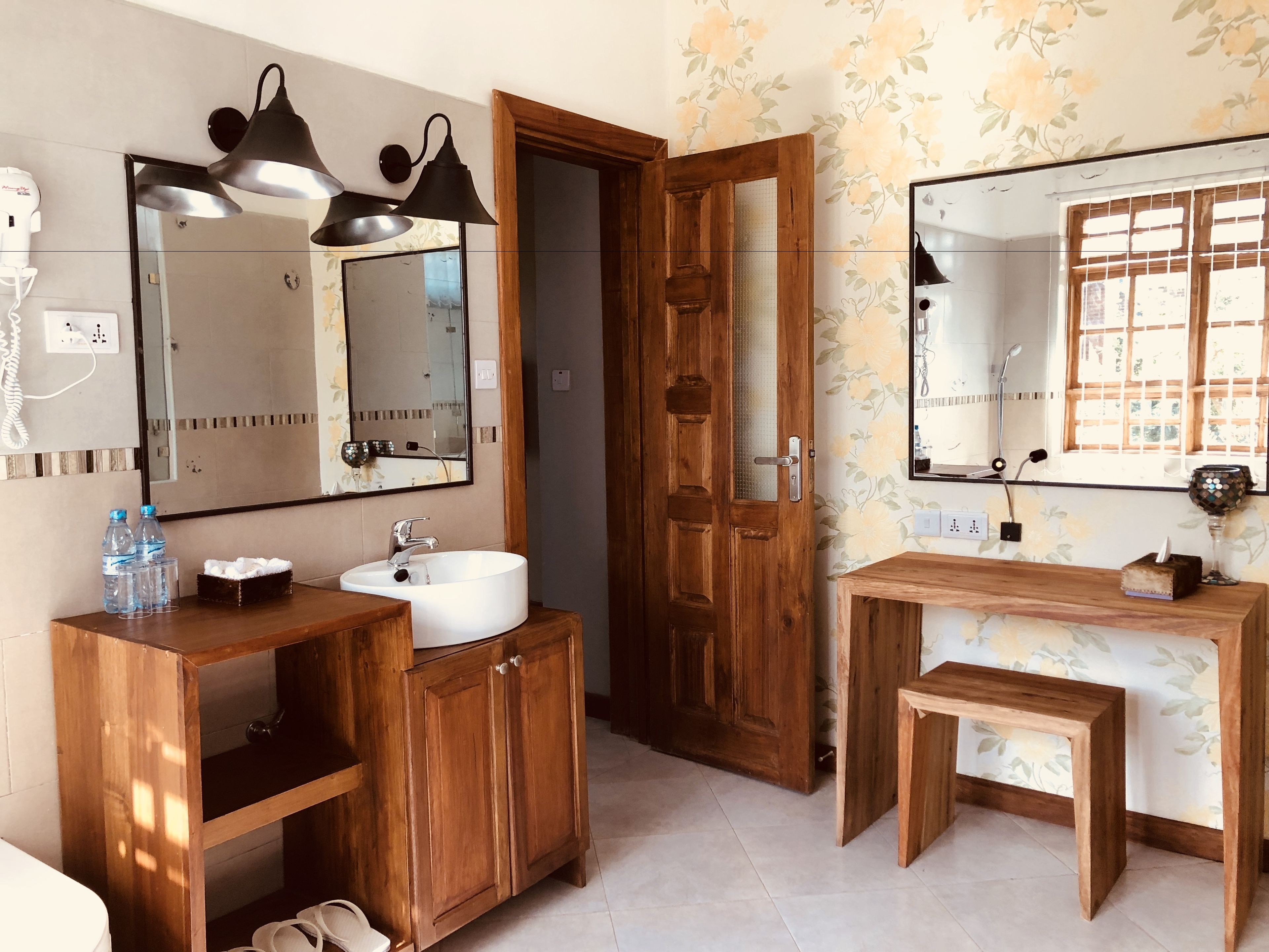 A bathroom with a wooden vanity, a sink, and a mirror. There are two hair dryers on the wall, a wooden door, and a window with a view.
