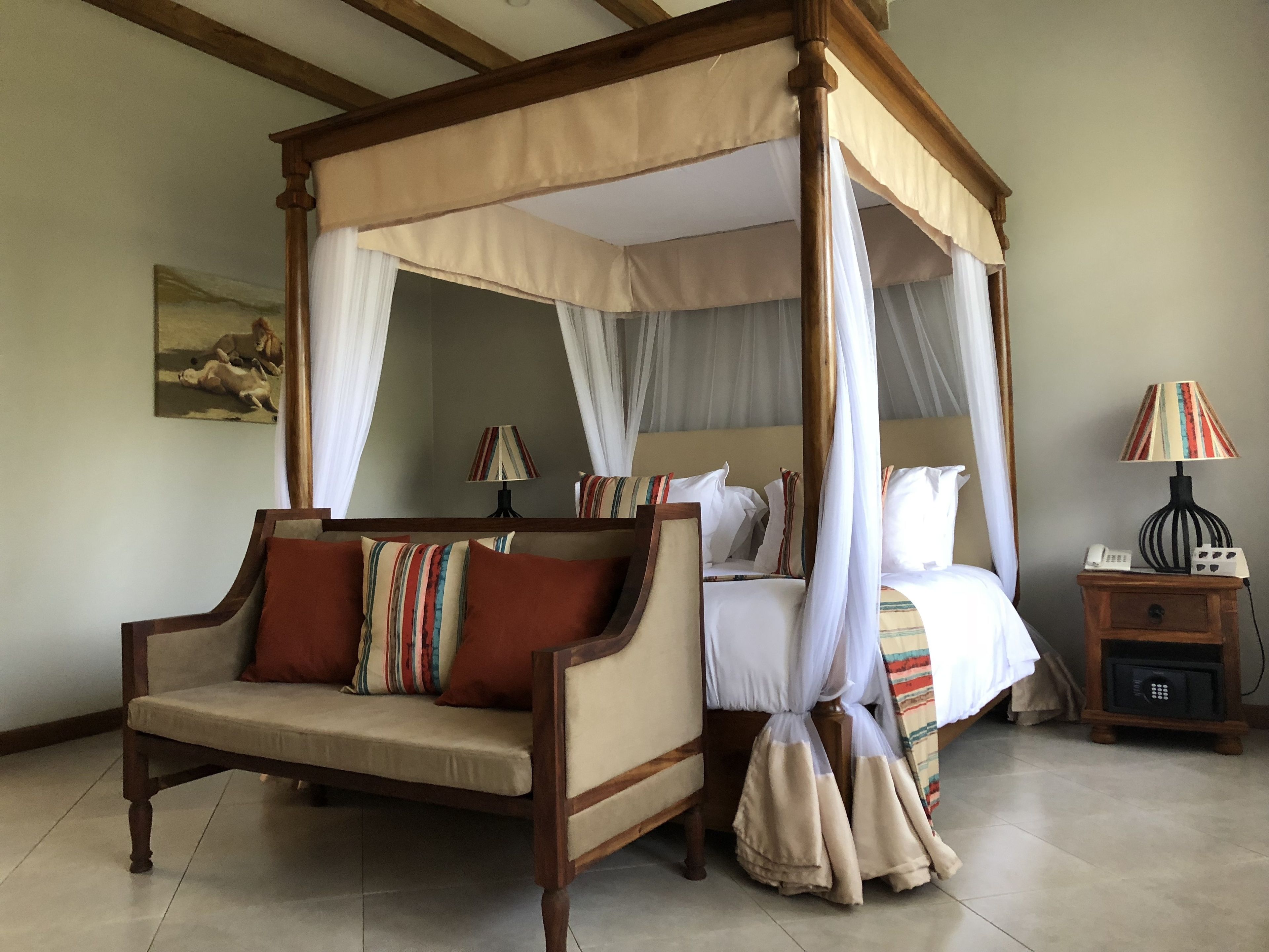A four-poster canopy bed with a wooden frame and white drapes, a sofa with striped cushions, a bedside table with a lamp, and a painting on the wall.
