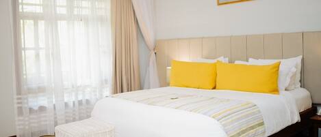 Un dormitorio con una cama, dos mesitas de noche, una mesita auxiliar, una ventana con cortinas traslúcidas y un cuadro en la pared.