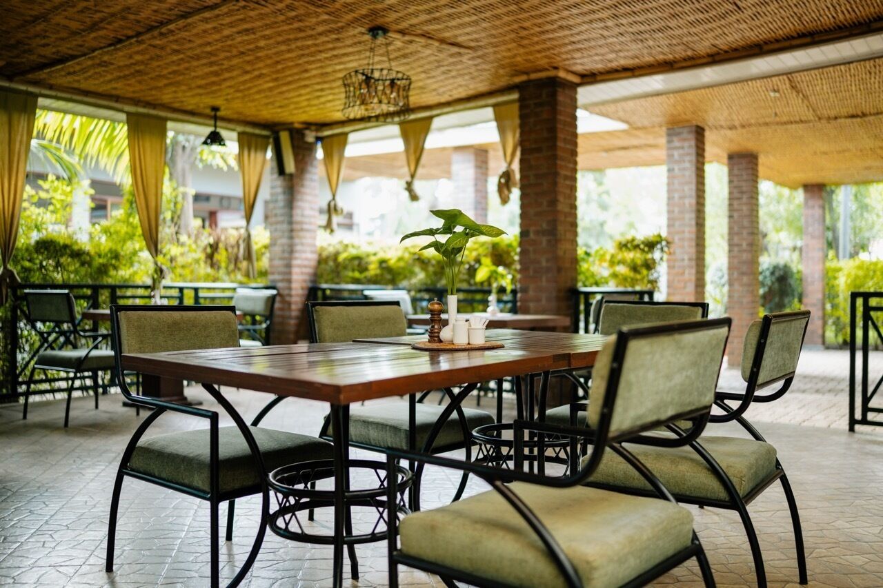 An outdoor dining area with a wooden table and chairs, surrounded by greenery and brick pillars.