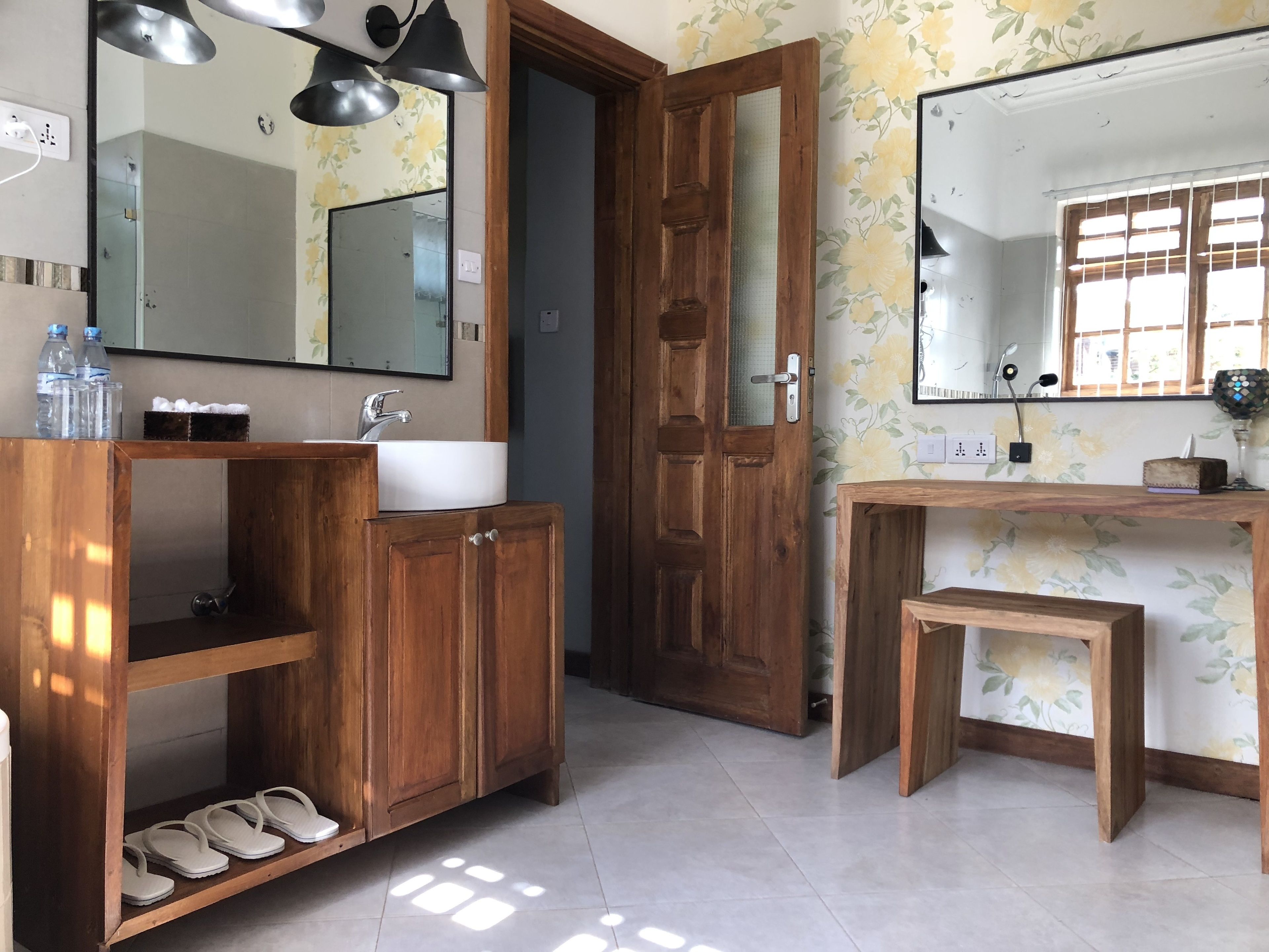 A bathroom with wooden cabinets, a sink, and a mirror. There are slippers under the cabinets and a small wooden bench.