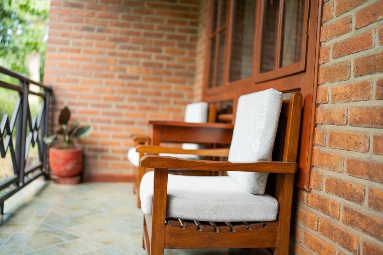 A wooden chair with a white cushion on a balcony with a brick wall and a window.