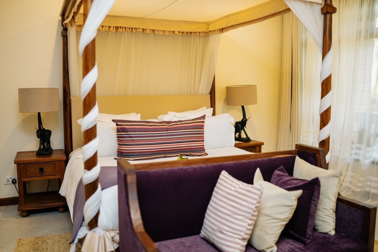 A four-poster canopy bed with a wooden frame, two bedside tables with lamps, a purple sofa with cushions, and a window with sheer curtains.