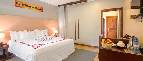 A hotel room with a large bed, a desk with a basket of fruit, a water bottle, and a mirror.
