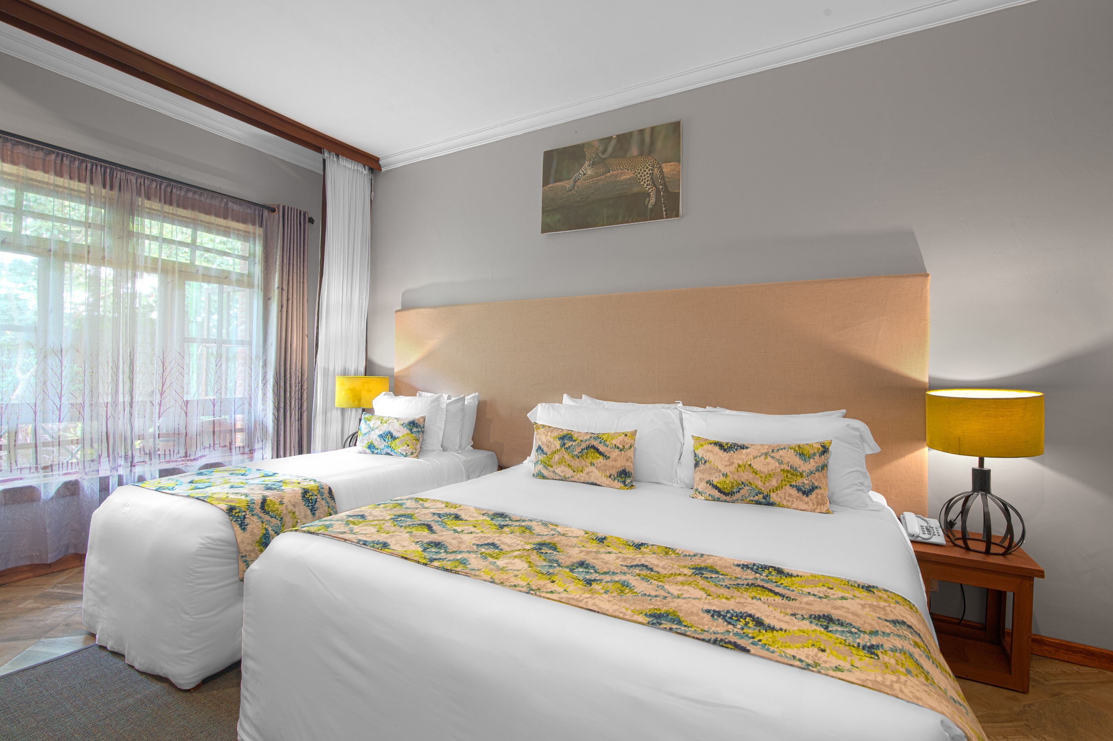 Una camera d'albergo con due letti, un'ampia finestra con tende, un quadro appeso al muro e un comodino con una lampada.
