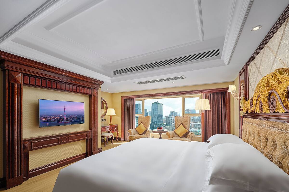 Una habitació d'hotel amb un llit gran, una televisió penjada a la paret, un escriptori amb una cadira i vista a un paisatge urbà per la finestra.