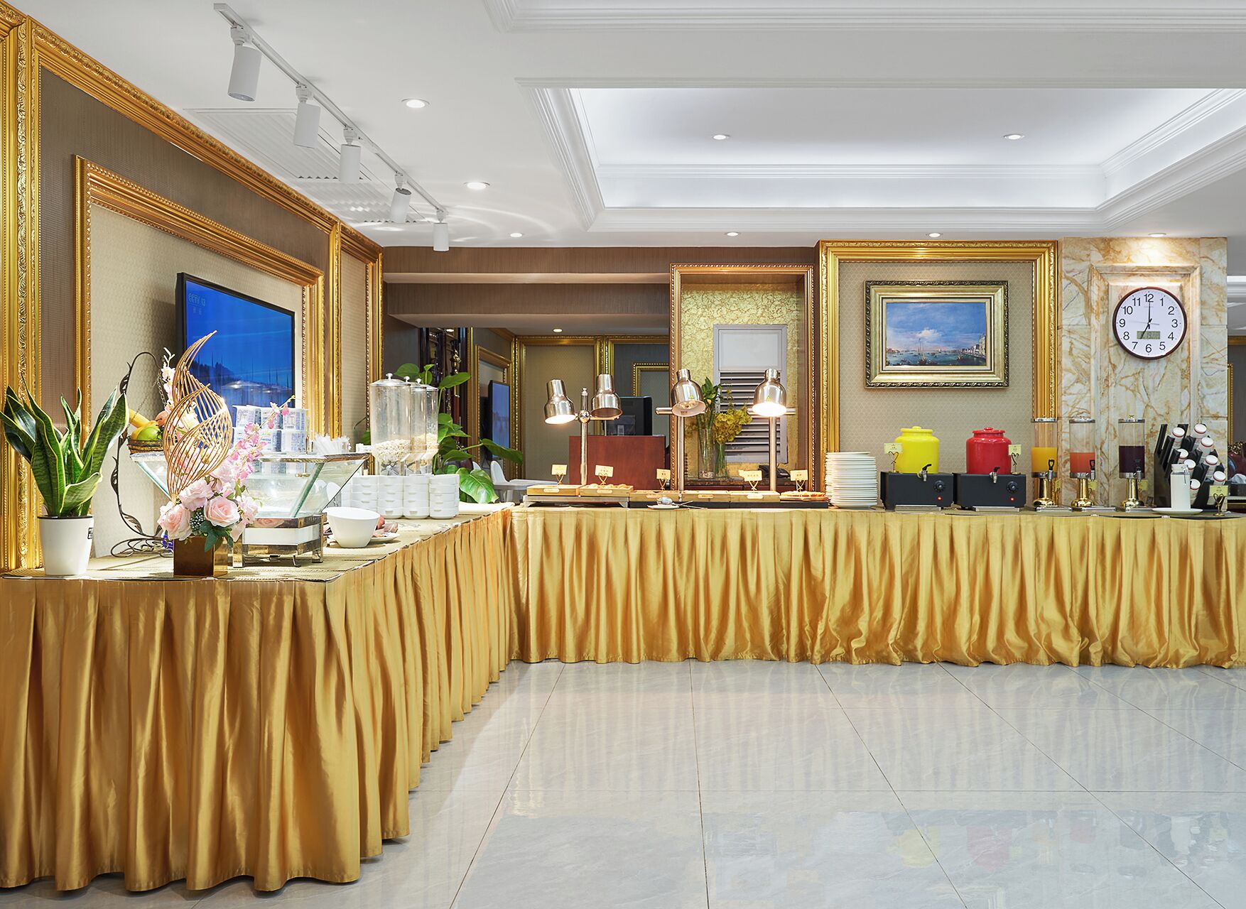 Elegante montaje de bufé con una mesa cubierta en tela dorada, diversas estaciones de comida y bebidas, y una decoración refinada que incluye un reloj grande y obras de arte enmarcadas.