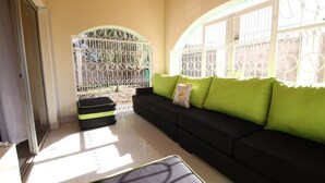 Habitación iluminada por el sol con un sofá negro y cojines verdes, una mesita y una vista a través de un portón blanco.