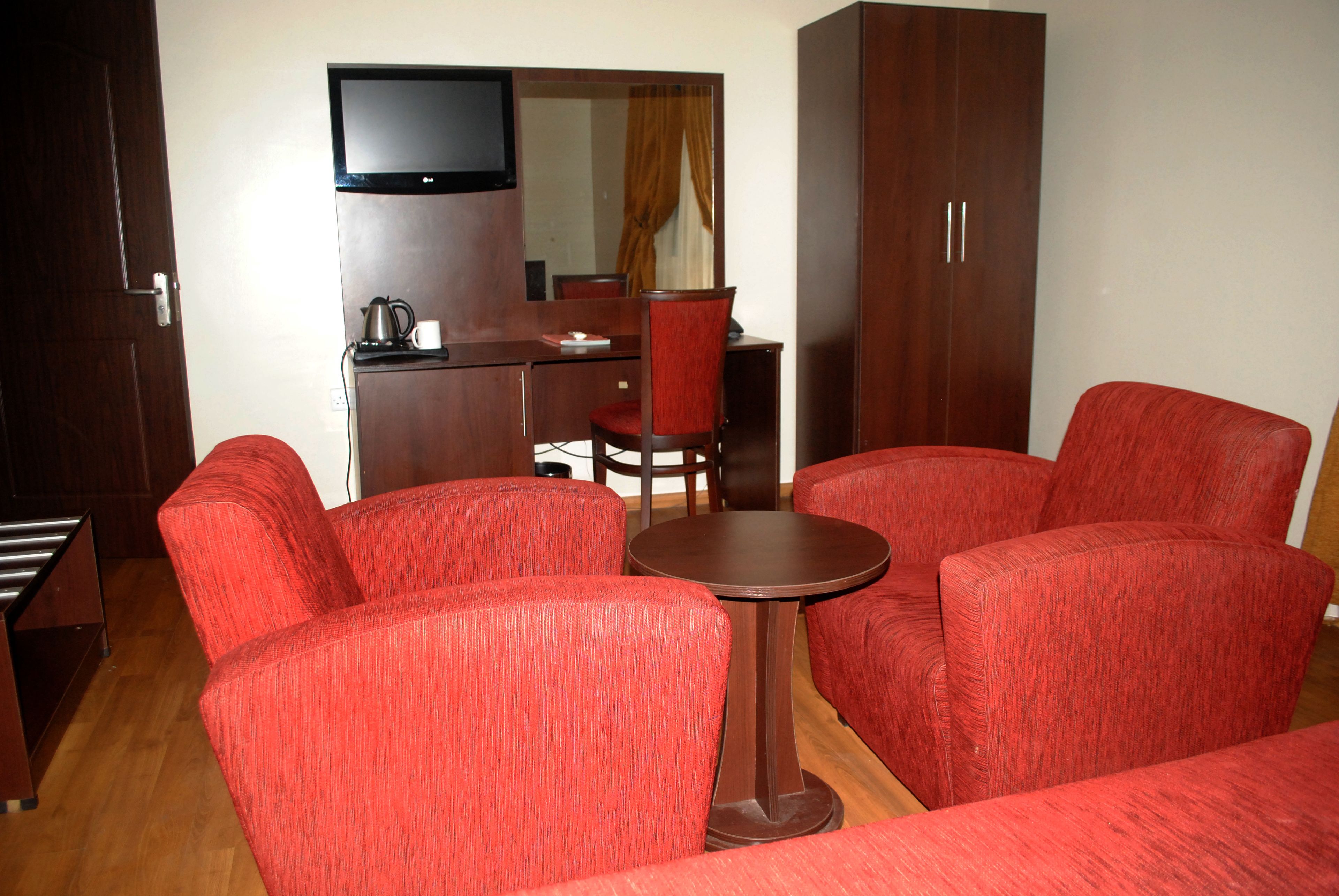 A hotel room with a red upholstered sofa set, a small round table, a television mounted on the wall, and a wooden wardrobe.
