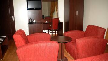 A hotel room with a red upholstered sofa set, a small round table, a television mounted on the wall, and a wooden wardrobe.