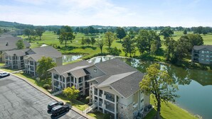 A multi-story hotel with a parking lot, a golf course in the background, and a body of water in the foreground.
