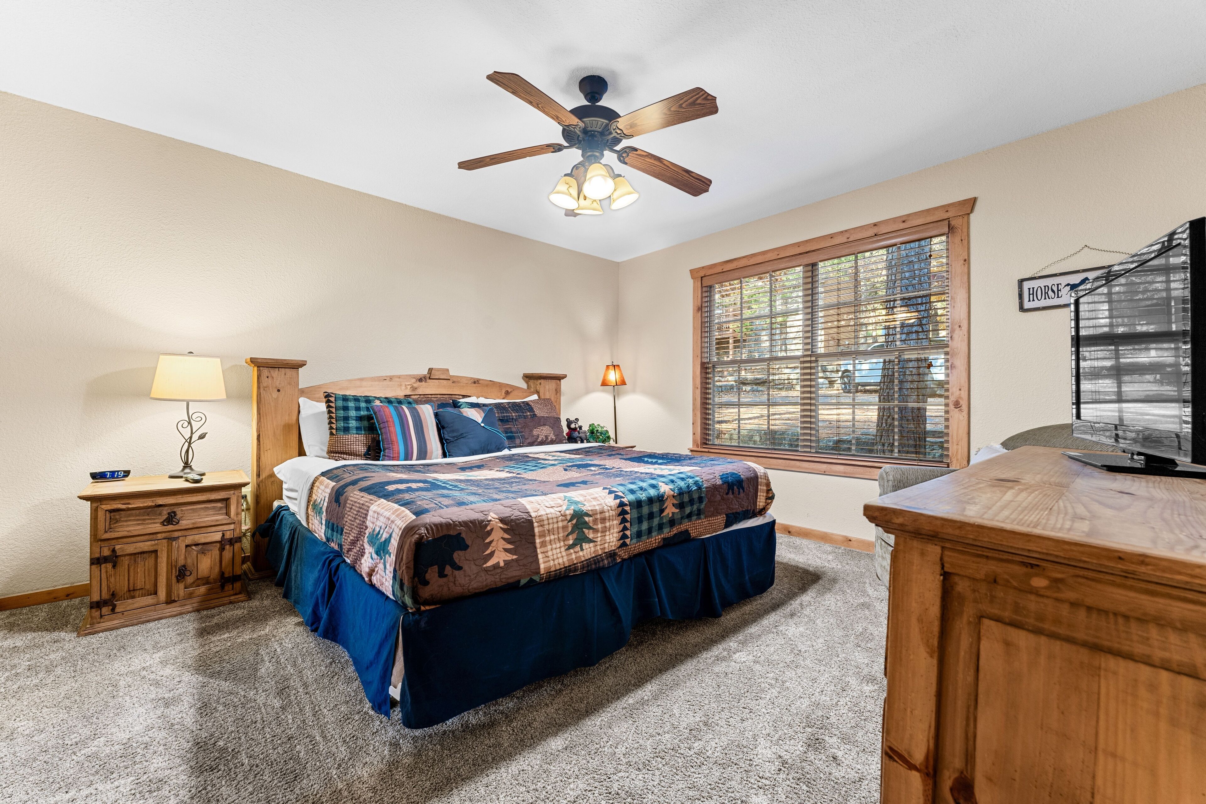 A bedroom with a bed, a wooden nightstand, a desk, a ceiling fan, and a window with blinds.