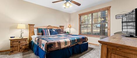 A bedroom with a bed, a wooden nightstand, a desk, a ceiling fan, and a window with blinds.