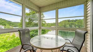A covered outdoor area with a round glass table and two chairs, overlooking a river and greenery. - Fort Taneycomo 2 Bedroom Condo by RedAwning (Branson)