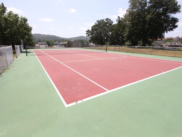 Un court de tennis en plein air, doté d’une surface verte et rouge, entouré d’une clôture en treillis métallique et d’arbres.