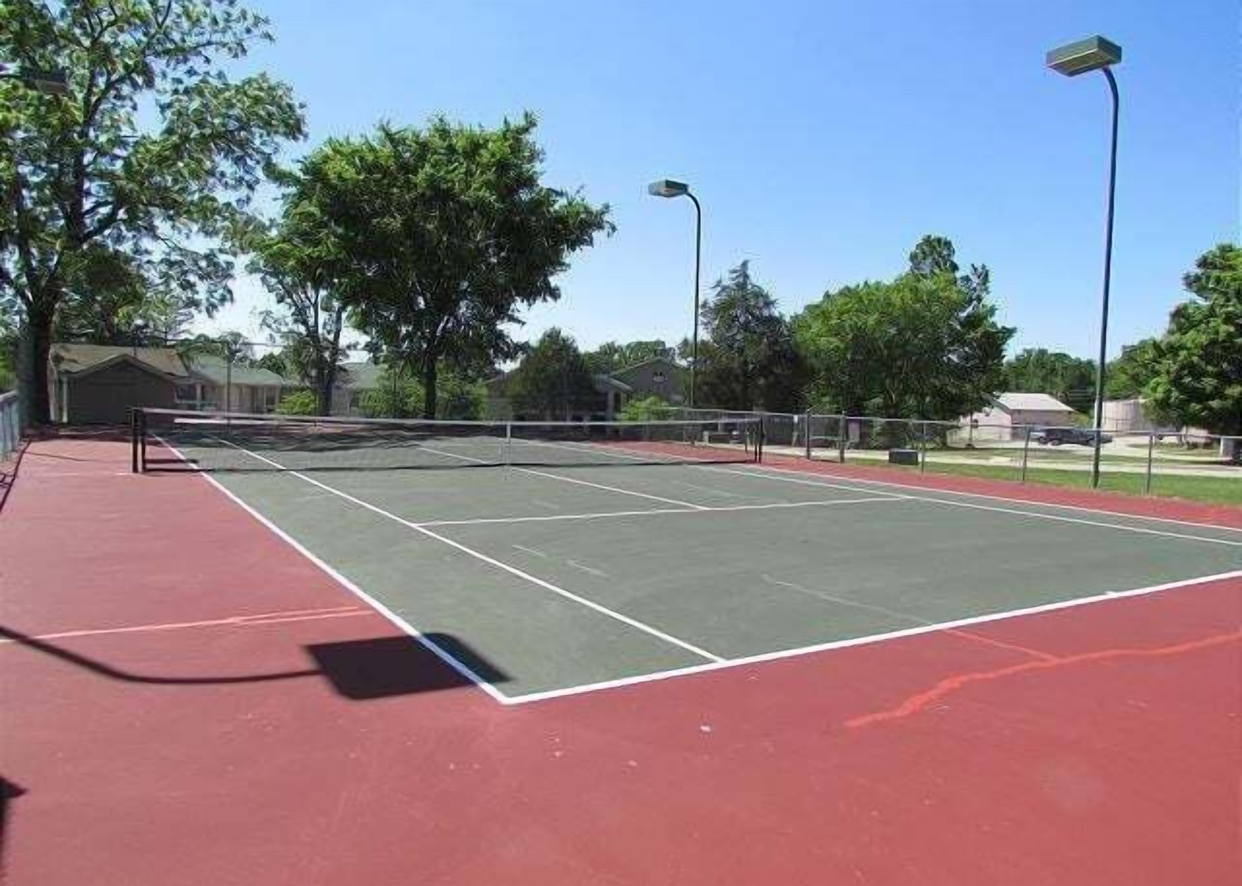 Un court de tennis extérieur doté d’une surface rouge, entouré d’arbres et d’une clôture.