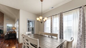 Un espace repas avec une table en bois et des chaises blanches, un lustre et de grandes fenêtres ornées de rideaux à motifs.
