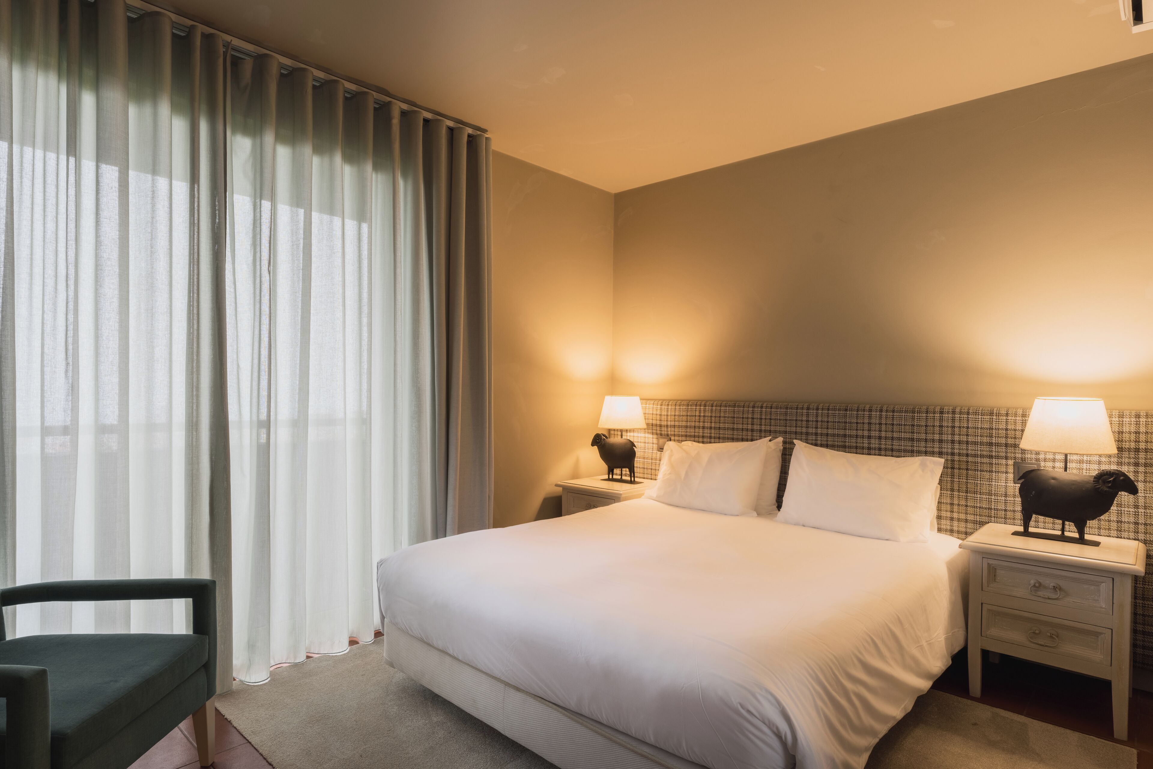 Une chambre d’hôtel avec un grand lit, deux tables de chevet avec des lampes, une chaise et des rideaux légers.