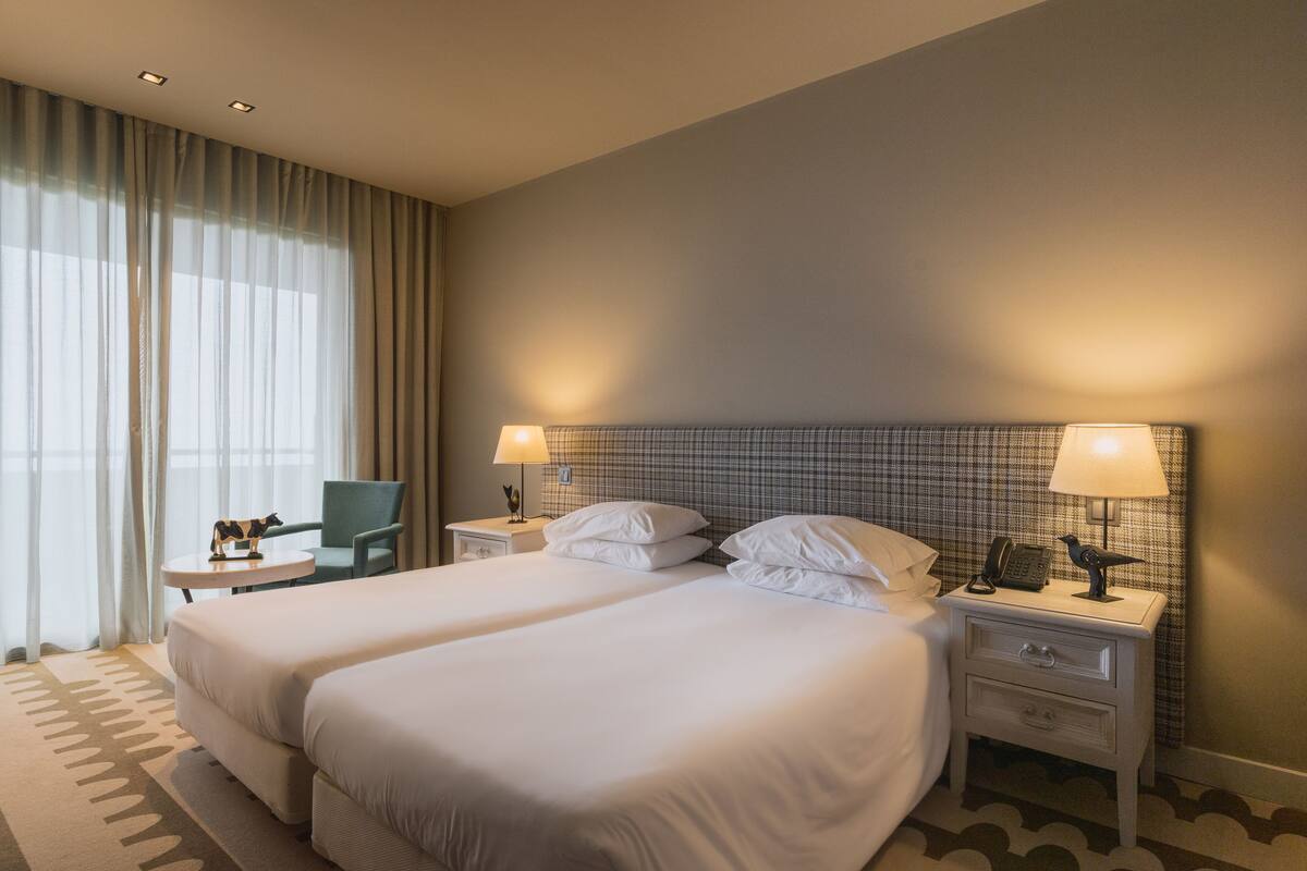 Hotellihuone, jossa on kaksi sänkyä, tuoli, yöpöytä lampun kanssa ja ikkuna, jossa on verhot.