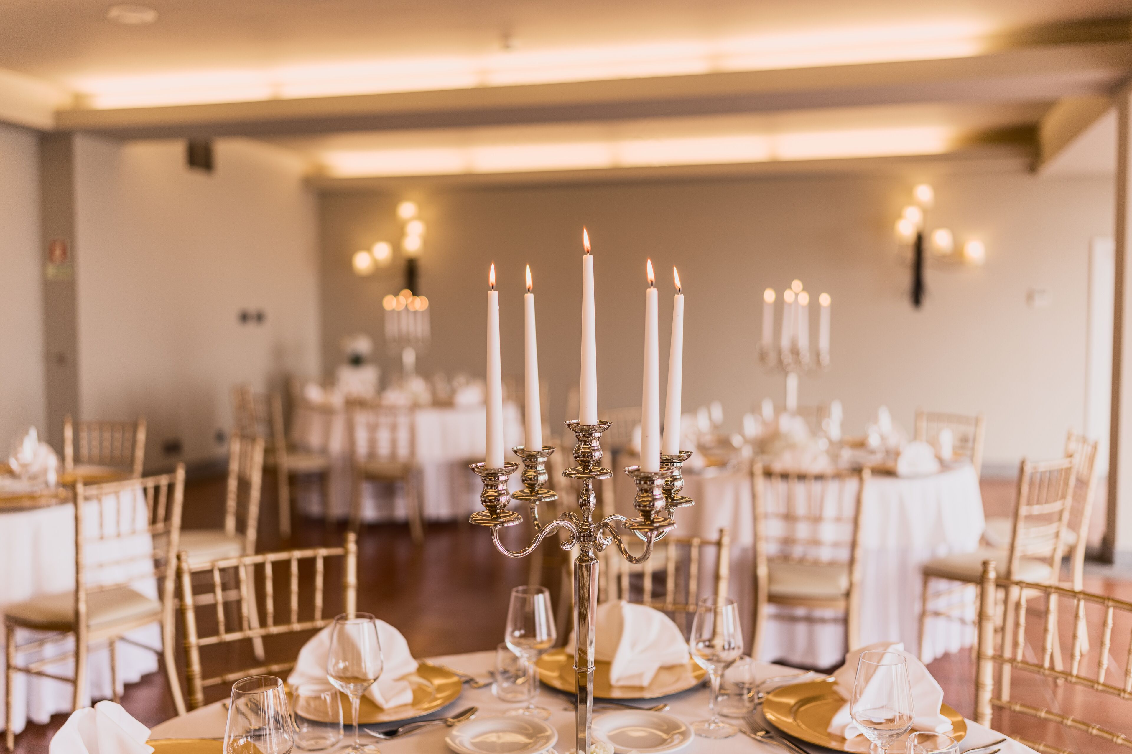 A dining area with tables set for a formal event, featuring white tablecloths, gold cutlery, and a central candelabra with lit candles.