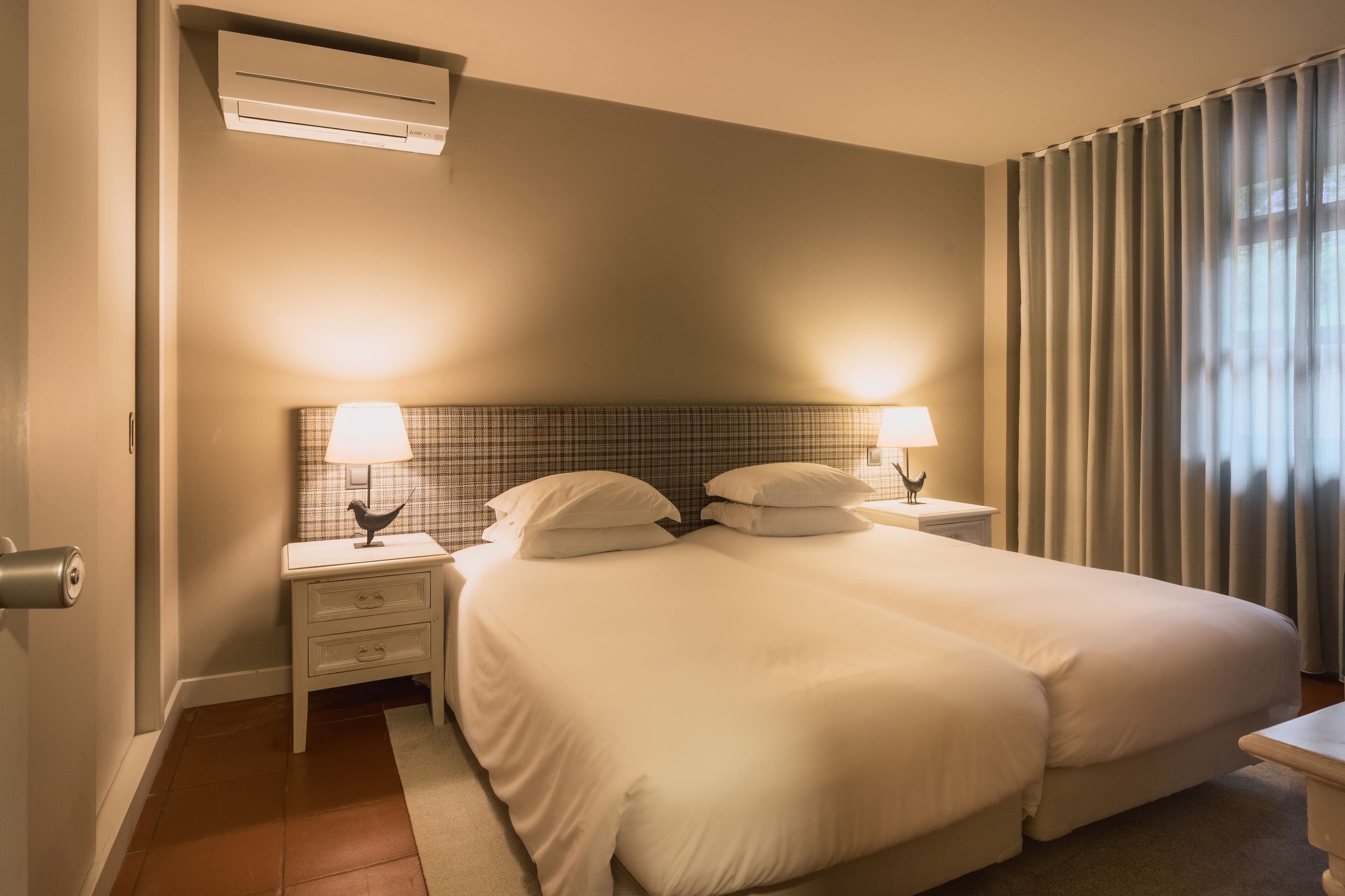 A hotel room with a large bed, two bedside tables with lamps, a wall-mounted air conditioner, and a window with curtains.