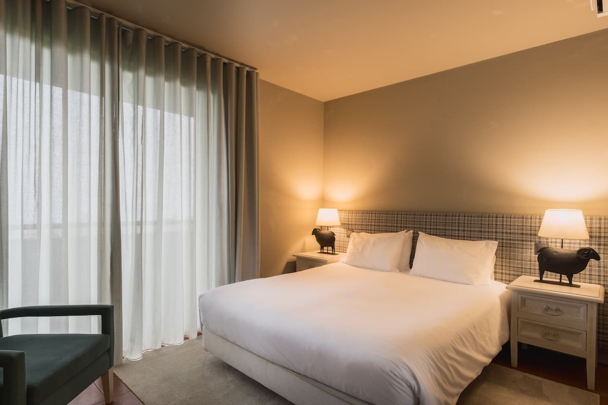 Hotellihuone, jossa on suuri sänky, kaksi yöpöytää, joissa on lamput, tuoli ja läpikuultavat verhot.