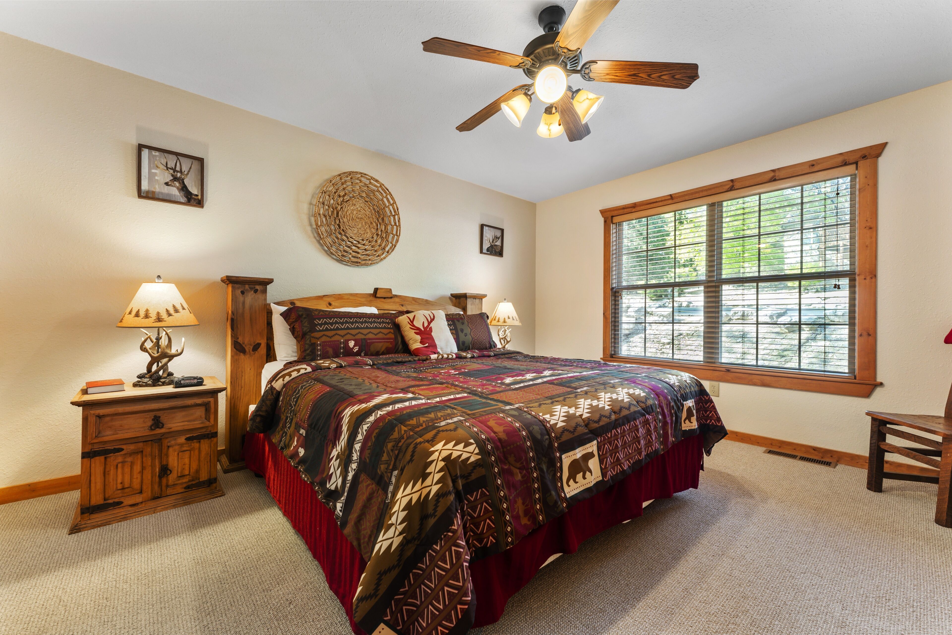 A bedroom with a large bed, a ceiling fan, a window with blinds, a nightstand, and a chair.