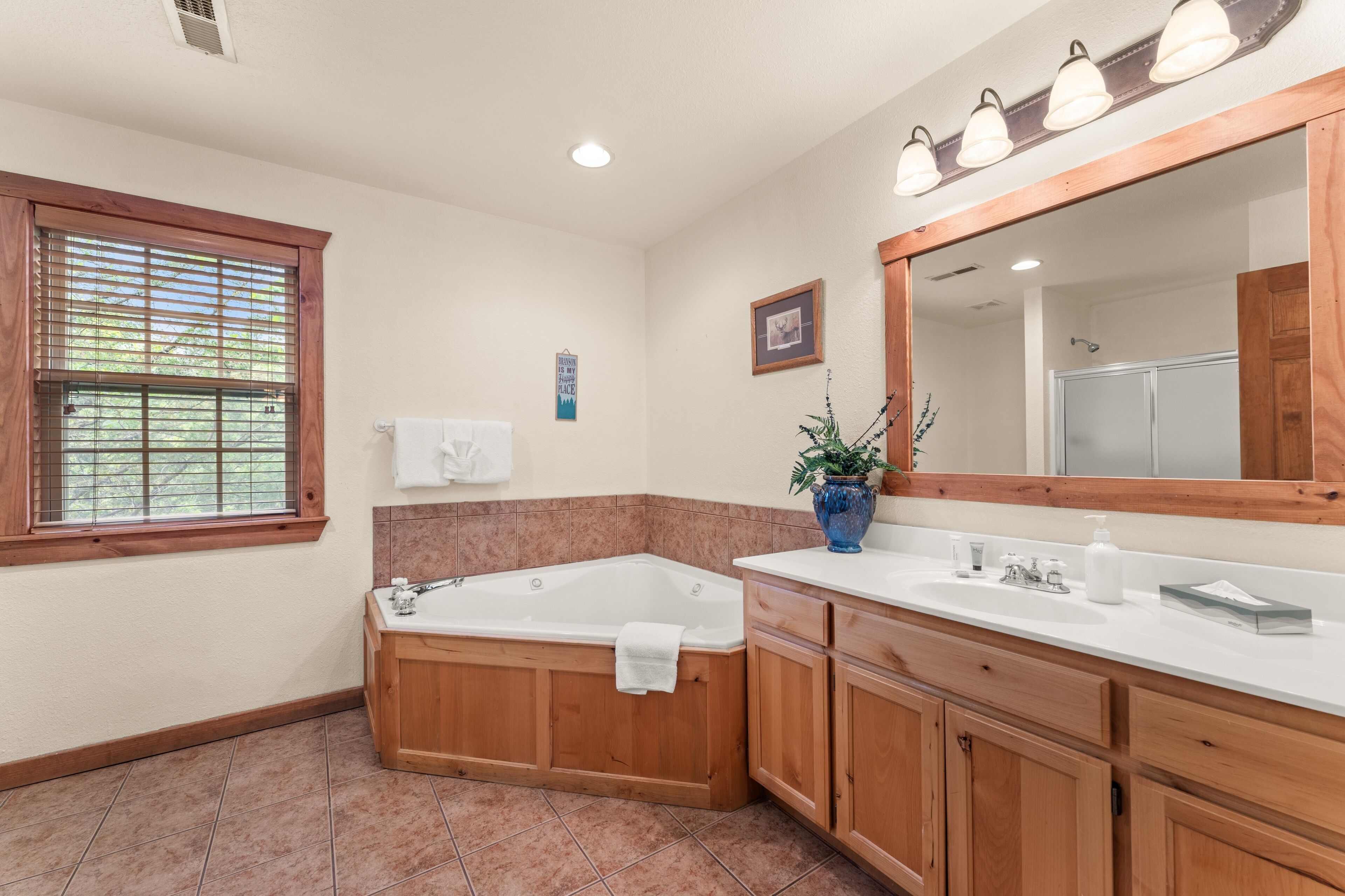 A bathroom with a bathtub, vanity, wooden cabinets, a large mirror, and a window with blinds.