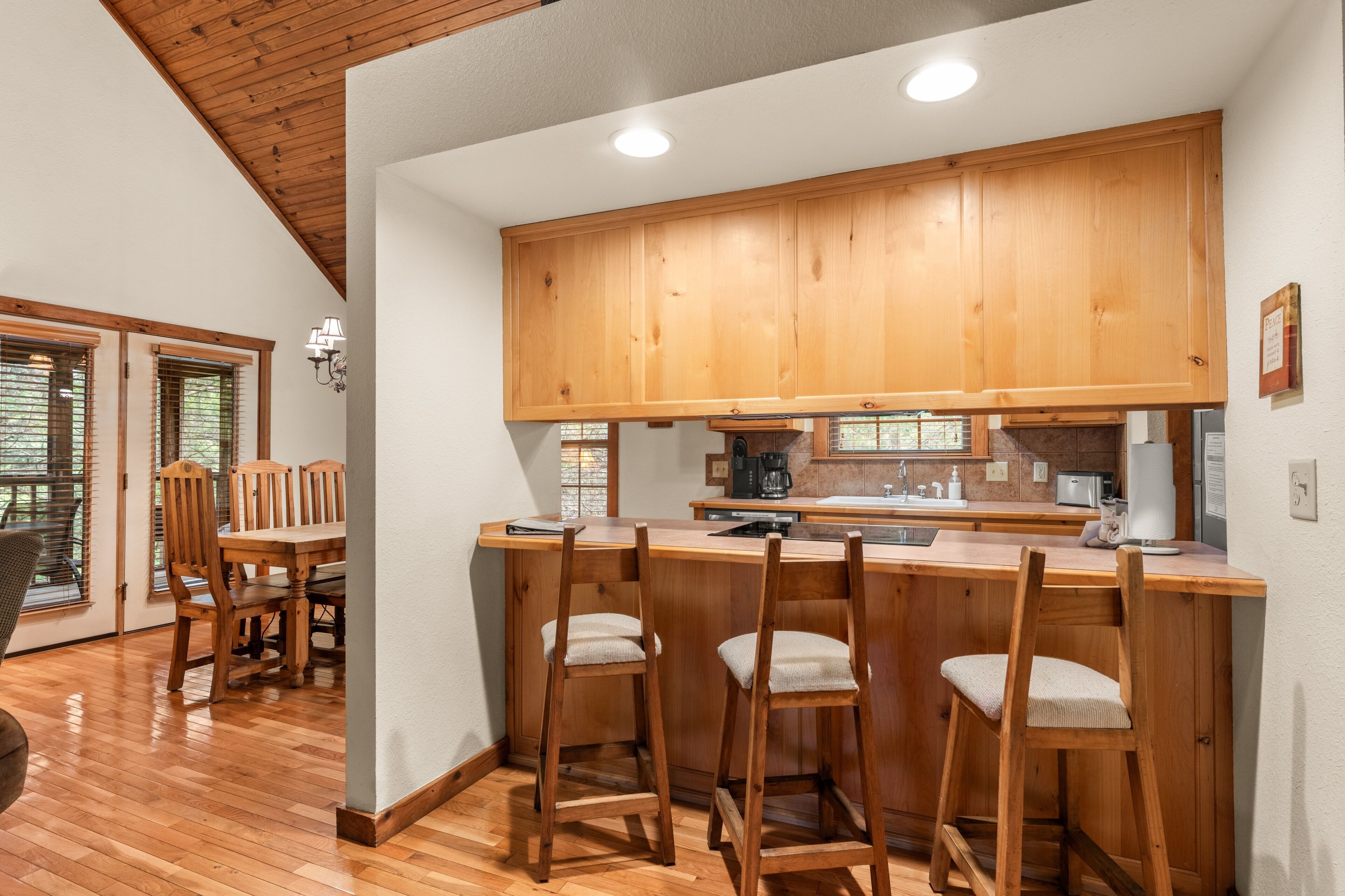 Une cuisine avec des meubles en bois, un coin bar avec des tabourets, une table à manger avec des chaises et une vue sur l’extérieur par une fenêtre.
